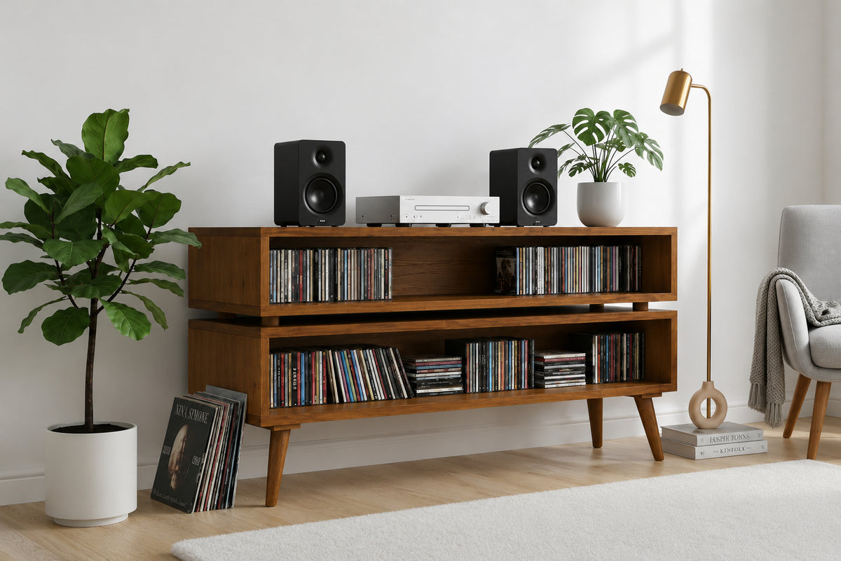 A mid-century modern wooden media console with open shelves and tapered legs, styled in a bright living room with CD cases, speakers, plants, a floor lamp, and a chair.