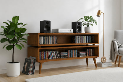 A mid-century modern wooden media console with open shelves and tapered legs, styled in a bright living room with CD cases, speakers, plants, a floor lamp, and a chair.