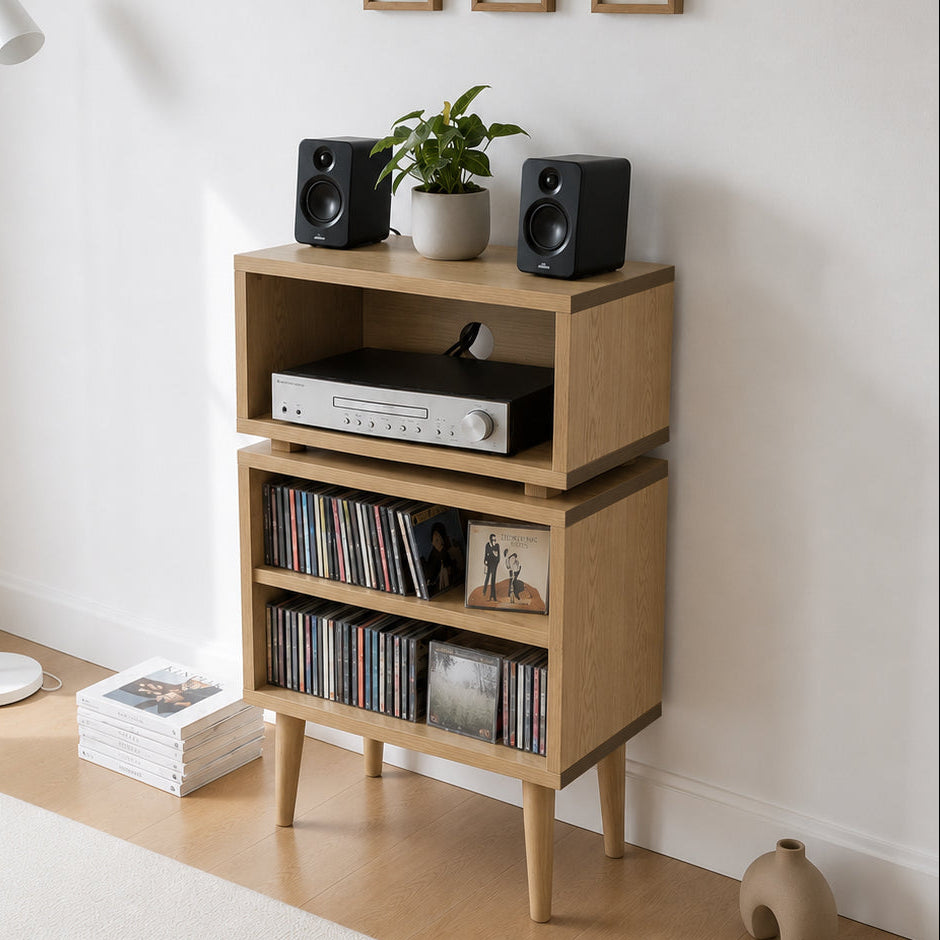 A light natural wood three-tier media cabinet with tapered legs, styled in a minimalist room corner with a white floor lamp, framed wall art, speakers, a plant, and a vinyl record player.