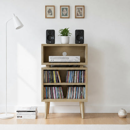 A light wood media console with three tiers and tapered legs, styled with speakers, a small plant, a DVD player, and records, set against a white wall in a minimalist room.